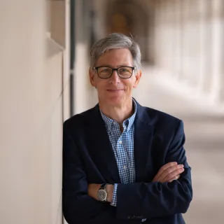 "Peter Kastor leans against a wall and smiles at the camera. He is wearing dark-brimmed glasses and a suit."