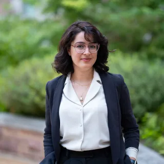 Image of Marjan Wardaki - she is wearing a white button-up shirt with a black long-sleeve blazer. She has wide-brimmed glasses. Wardaki smiles at the camera.